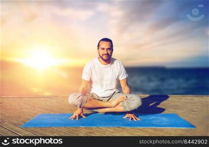 fitness, yoga, people and healthy lifestyle concept - man doing scale pose lotus variation on mat outdoors on wooden pier over sea background. man doing yoga scale pose outdoors