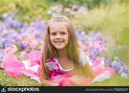 Five-year girl in a beautiful dress in a bed of flowers. Happy six year old girl in a lush evening pink dress walking through the green garden