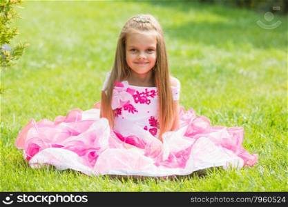 Five-year girl in a beautiful dress sat on the lawn in the green garden. Happy six year old girl in a lush evening pink dress walking through the green garden