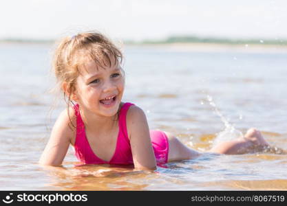 Five-year girl lies in water in the shallows of the river, laughing and looking to the left