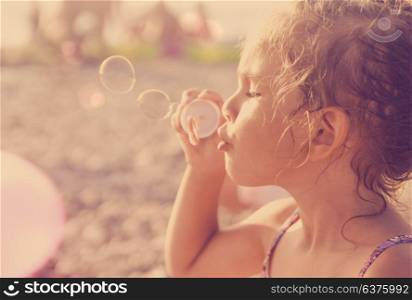 Five year old caucasian beautiful little girl blows soap bubbles on a summer beach on a sunny hot day. Happy carefree childhood. Toned orange purple effect.