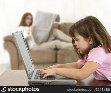 Five year old girl laying on table with laptop while mom (out of focus) reads newspaper on couch. Shot in studio.