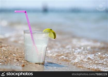 Fizzy drink or water with straw and lemon slice on glass standing on beach by sea. Summer vacation