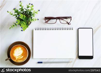 Flat lay business office accessories on modern white marble table, blank page of notebook, mobile with empty white screen, coffee cup, glasses, pencil and green plant pot.
