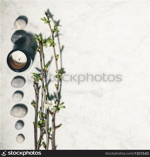 Flat lay composition with beautiful spring cherry branches, natural candle and grey stones on white marble background. SPA, yoga, relaxation, natural cosmetics, zen like concept