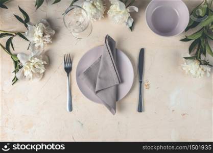 Flat lay composition with white peony flowers, empty plate with napkin and cutlery on a light pink concrete background