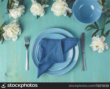 Flat lay composition with white peony flowers, empty plate with napkin and cutlery on wooden turquoise table surface, festive flat lay arrangement.