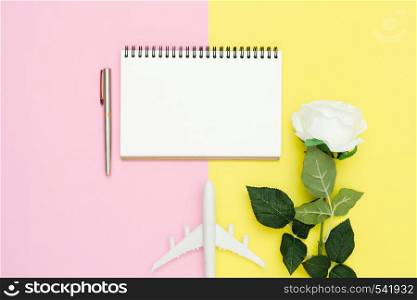 Flat lay design of travel summer concept - Top view mockup of white paper notebook, stationary, plane and white rose on pink and yellow pastel color screen. Travel in summer on pastel color background