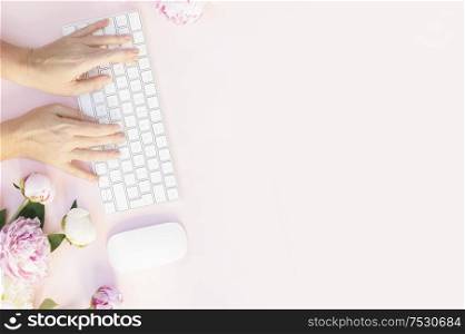 Flat lay home office workspace - modern keyboard with peony flowers and someones two hands typing, copy space on pink background. Top view home office workspace