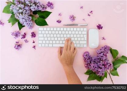 Flat lay home office workspace - modern keyboard with someone hand typing, fresh lilac flowers. Top view home office workspace