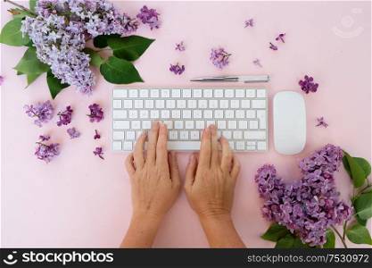 Flat lay home office workspace - modern keyboard with two hands typing, fresh lilac flowers. Top view home office workspace