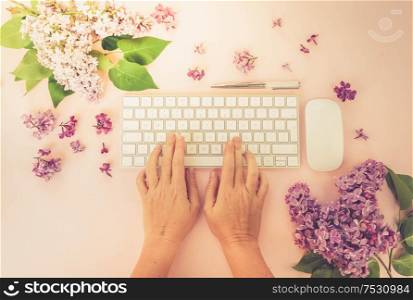 Flat lay home office workspace - modern keyboard with two hands typing, fresh lilac flowers, toned. Top view home office workspace