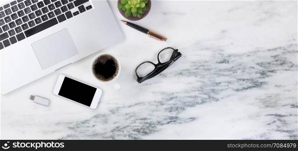 Flat lay of a partial laptop, baby plant, pen, reading glasses, smartphone, thumb drive and coffee on natural marble desktop