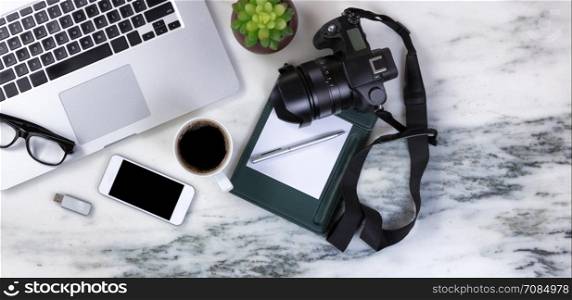 Flat lay of a partial laptop, camera, baby plant, pen, reading glasses, smartphone, thumb drive and coffee on natural marble desktop