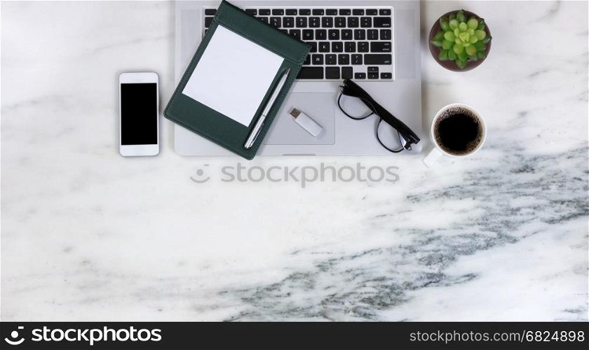 Flat lay of a partial laptop, plant, smartphone, pen, paper, reading glasses, thumb drive, and dark coffee on natural marble desktop