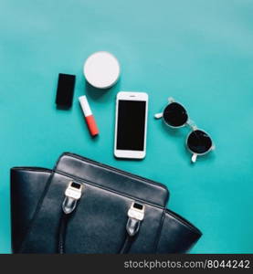 Flat lay of black leather woman bag open out with cosmetics, accessories and smartphone on green background