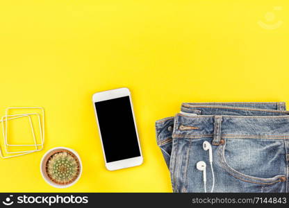 Flat lay of blue jeans, cactus and smartphone headphones on bold yellow paper background with copy space. Overhead view of woman casual outfit. Trendy hipster look top view
