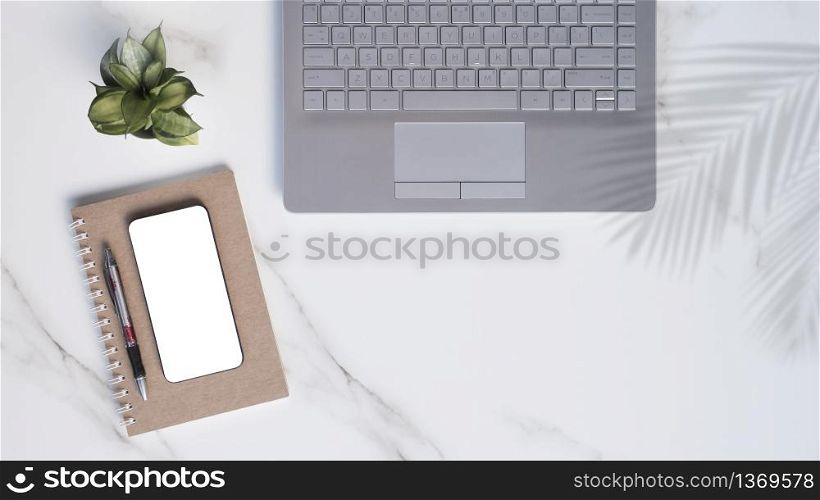 Flat lay of modern white marble office desk table with laptop, smartphone mock up, ballpoint pen, spiral notebook with little green houseplant and palm leaves shadow on tabletop