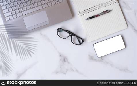 Flat lay of modern white marble office desk table with laptop, smartphone mock up, eyeglasses, ballpoint pen, spiral notebook with palm leaves shadow on tabletop