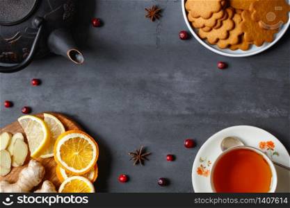 Flat lay of natural remedies for colds and flu, as well as for cozy tea drinking in cold weather, such as tea, lemon, ginger, dried oranges, anise, Gingerbread Cookie on black stone background.