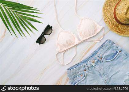 Flat lay of summer items with pastel bikini, sunglasses, jeans, hat and green tropical plant on marble background, fashion and summer concept