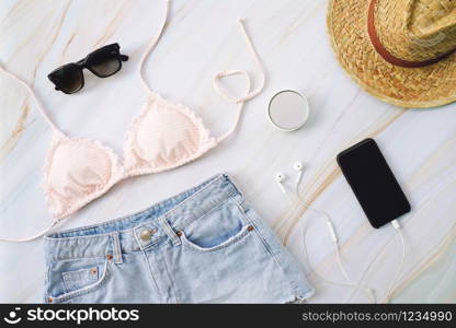 Flat lay of summer items with pastel bikini, sunglasses, jeans, hat, makeup, hat and smartphone on marble background, fashion and summer concept