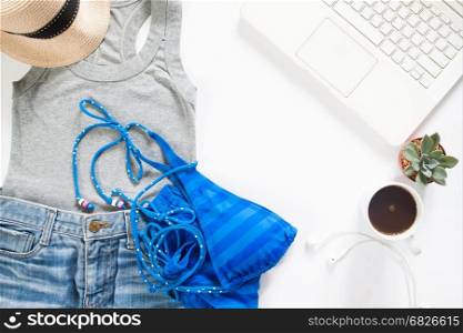 Flat lay of summertime clothing with blue stripe bikini, laptop and cup of coffee on white background