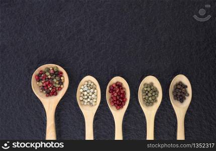 Flat lay of various colors peppercorns in wooden spoon on black stone slate background