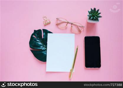 Flat lay of workspace desk, minimal stationery with smartphone and notebook, paper, pen, clip and eyeglasses on pink color background with copy space