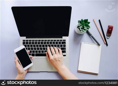 Flat lay of workspace desk with woman hands typing on laptop and using smartphone with note paper and stationery on purple background, business and mockup concept
