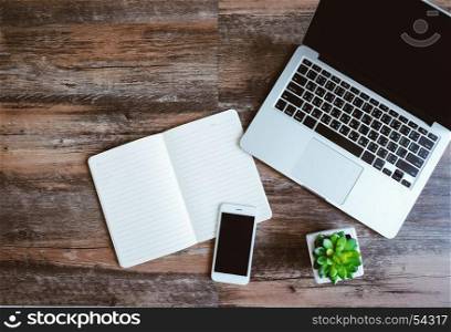 Flat lay photo of workspace desk with laptop, smartphone, blank notebook and green plant with copy space wooden background