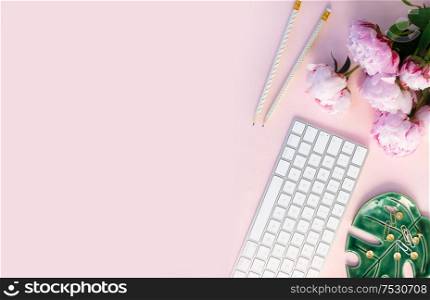 Flat lay top view home office workspace - modern keyboard with female accessories and peony flowers, copy space on pink desk background. Top view home office workspace