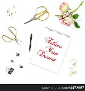 Flat lay with sketchbook, rose flowers, golden accessories on white background. Motivation quote Follow Your Dreams