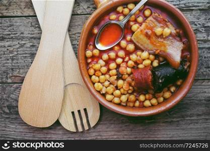 Flat of chickpeas, sausage and bacon in a crockpot by wooden spoon and fork. Typical food from Madrid, Spain, with a rustic wooden board as a background.