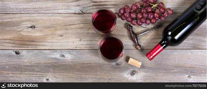 Flat view of a bottle of red wine, antique corkscrew, grapes, and drinking glasses on rustic wooden boards
