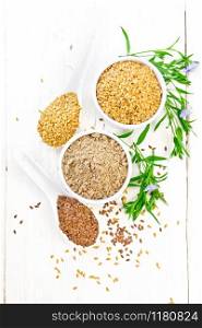 Flaxseed flour in a bowl, white and brown linen seeds in two spoons and on table, leaves and blue flax flowers on background of light wooden board from above