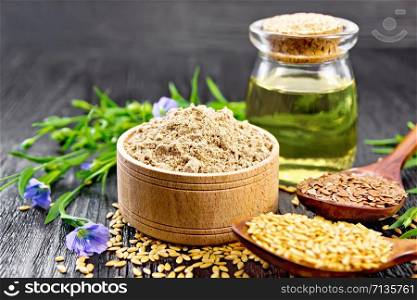 Flaxseed flour in a bowl, white and brown seeds in spoons and on table, flax leaves and flowers, oil in a glass jar on dark wooden board background