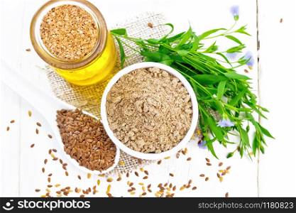 Flaxseed flour in bowl, linen seeds in a spoon, oil in a glass jar on burlap napkin, leaves and flowers of flax on wooden board background from above