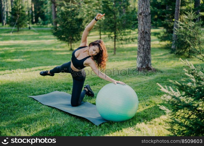 Flexible active woman in sportsclothes makes fitness exercises on karemat with fitball, raises arms and breathes fresh air in forest has morning workout poses over nature background. Healthy lifestyle