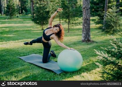 Flexible active woman in sportsclothes makes fitness exercises on karemat with fitball, raises arms and breathes fresh air in forest has morning workout poses over nature background. Healthy lifestyle