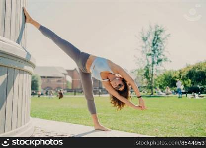 Flexible slim woman does stretching exercises outdoor stays in good physical shape dressed in cropped top and leggings stands on one leg leans with arms raised warms up before cardio training