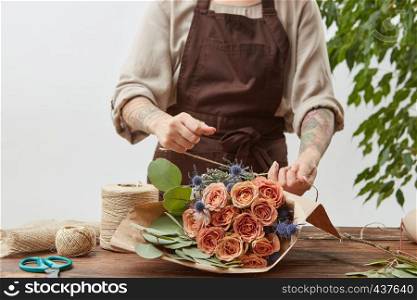 Floral design studio, woman's hands are making decorative bouquet from fresh natural fragrant flowers roses living coral color on a light background . Concept of flower shop.. Florist workplace - woman's hands are arranging a new bouquet with roses and decorative green leaves on a wooden table, process step by step. Place for text. Mother's Day.