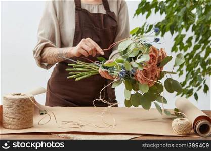 Floral design studio, woman's hands with tatto are making decorative bouquet from fresh natural fragrant flowers roses living coral color on a gray background . Concept of flower shop.. Female florist is decorating beautiful bouquet from fresh natural roses step by step at the table with paper and rope on it. Mother's Day holiday.
