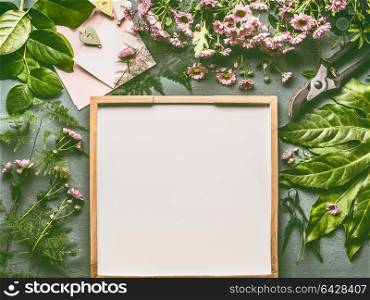 Florist desktop with a lot of decorative fresh green leaves , pink flowers and greeting card mock up around withe tray for florist work, top view, flat lay, frame with copy space