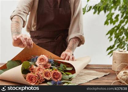 Florist woman in a brown apron is wrapping fresh rose bouquet in a decorative paper on a wooden table on alight background. Process step by step. Concept of floral shop.. Gardener girl make bouquet for a holiday from living coral roses and branches of green leaf on a wooden table on a light background. Mother's Day.