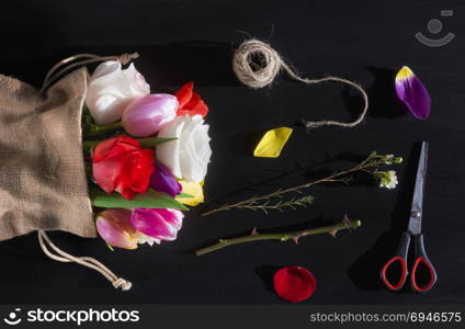 Florist workplace with a bouquet of roses and tulips, wrapped in a jute sack, surrounded by petals, scissors and a clew of linen, on a black table.