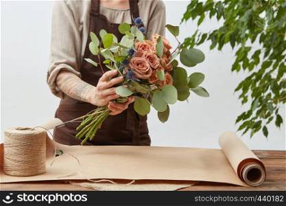 Florist workplace - woman's hands are arranging a new bouquet with roses, decorative green leaves on a light background step by step. Place for text.. Young woman florist is making bouquet with fresh flowers roses living coral color at the table with paper and rope. Process step by step. Concept floral shop.