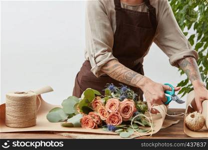 Florist workplace - woman's hands with tatto are arranging a new bouquet with roses, decorative green leaves on a gray background step by step. Place for text. Concept of flower shop.. Female florist is cutting paper for creating beautiful bouquet on a background of light wall. Place for text. Process of making bouquet step by step.