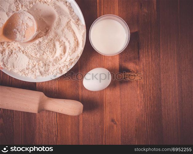 Flour in a bowl, white eggs and a wooden rolling pin on a wooden background. Top view