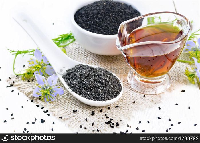 Flour Nigella sativa in a spoon, black cumin seeds in a bowl and oil in gravy boat on burlap, sprigs of kalingi with blue flowers and leaves on light wooden board background